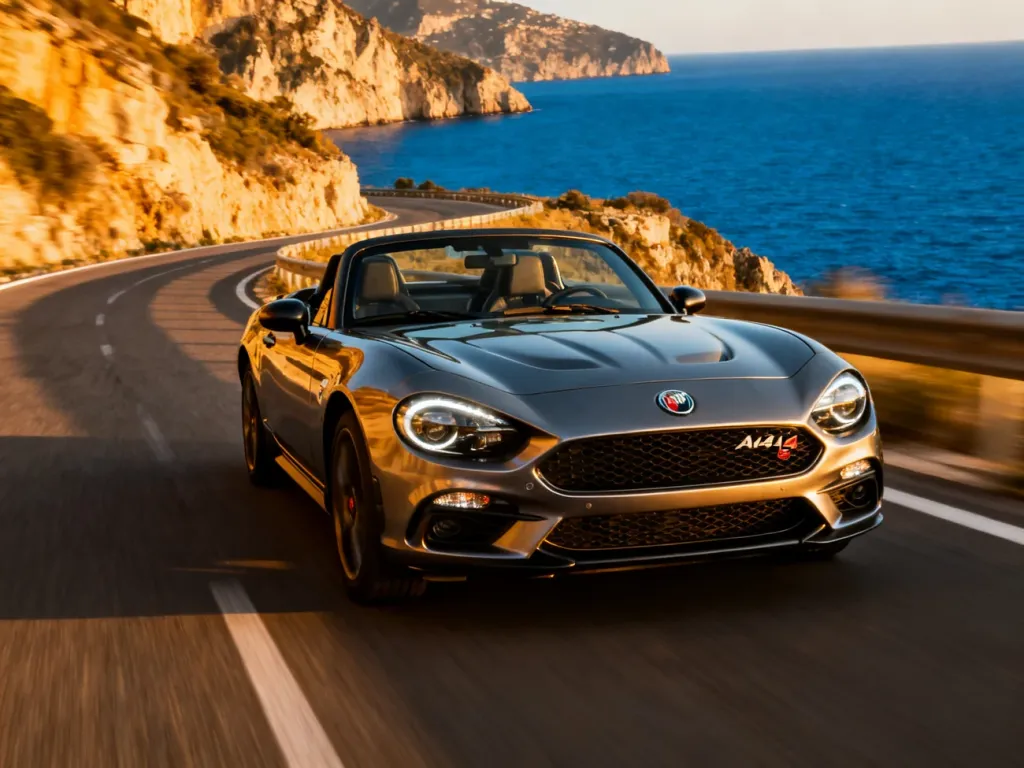 Sportowy Abarth 124 Spider pędzący krętą nadmorską drogą w blasku zachodzącego słońca.